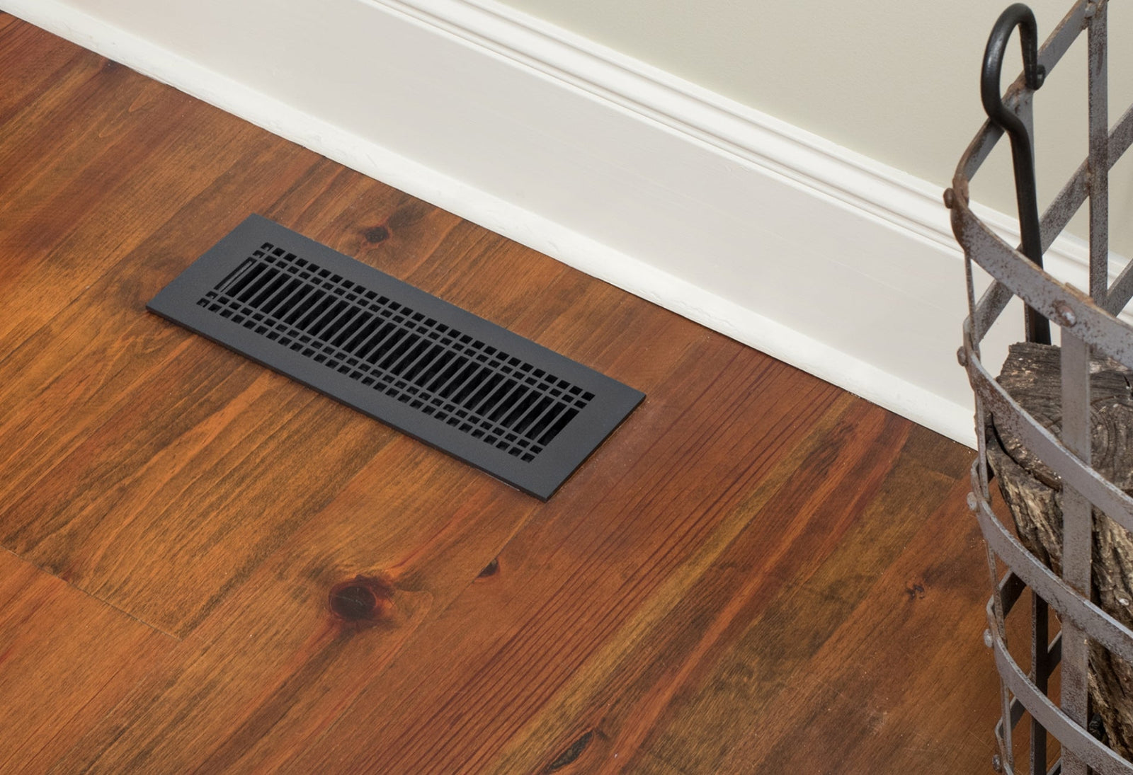 Close up of a black decorative register in a mid-tone wood floor against white and beige wall.